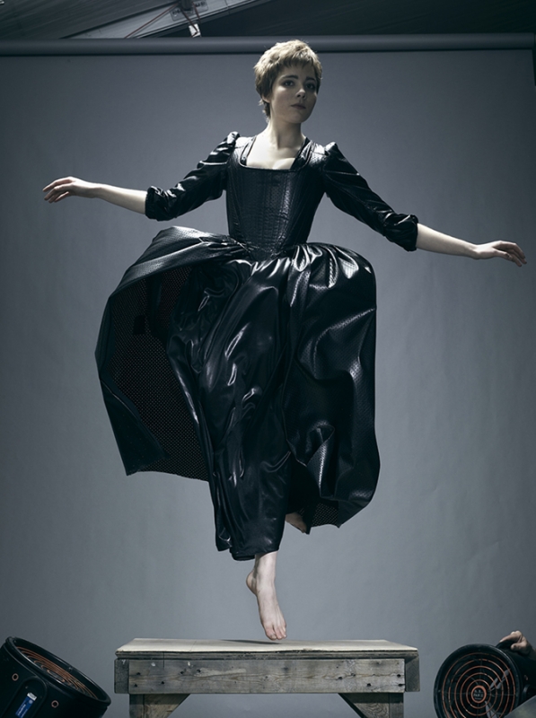 Woman in a black period dress leaps from a wooden platform in a studio setting. Her arms are outstretched and the dress is flared.