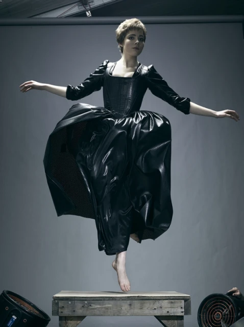 Woman in a black period dress leaps from a wooden platform in a studio setting. Her arms are outstretched and the dress is flared.