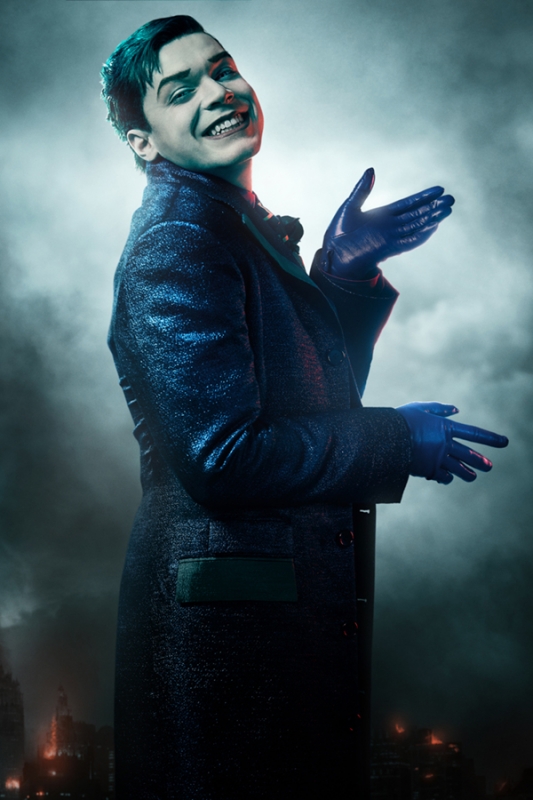 Portrait of a young adult male with a smirk, wearing a dark coat and blue gloves, against a blurred background of city lights and clouds.
