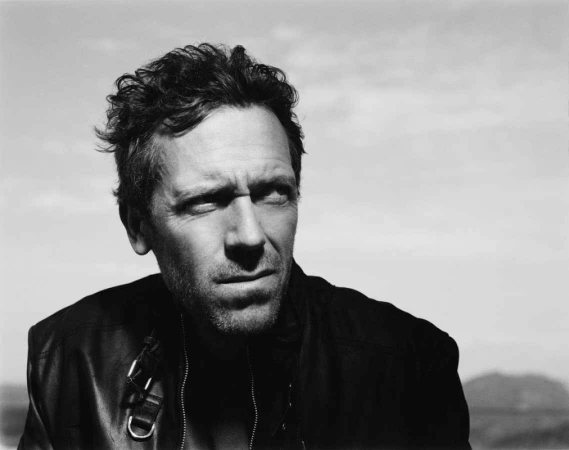 Black and white portrait of Hugh Laurie looking off-camera, wearing a dark jacket, set against a blurry outdoor background.