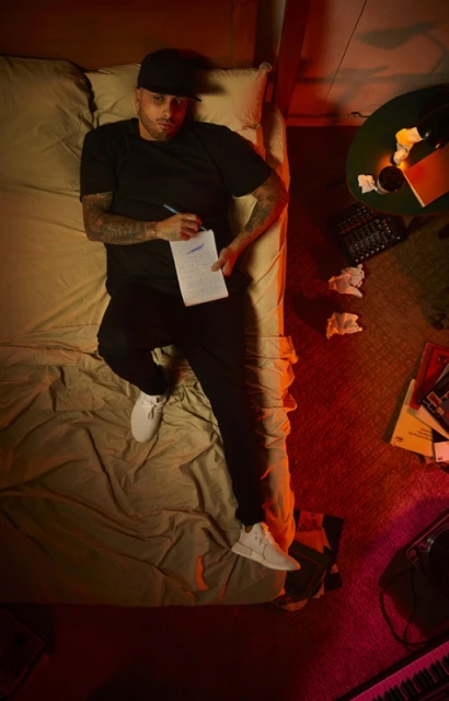 Overhead shot of a male musician writing on a notepad while lying on a bed in a room with music production equipment.