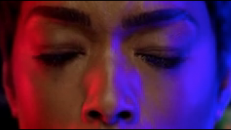 Close-up shot of a person's face with eyes closed, bathed in blue and red light, creating a dramatic and intimate portrait.