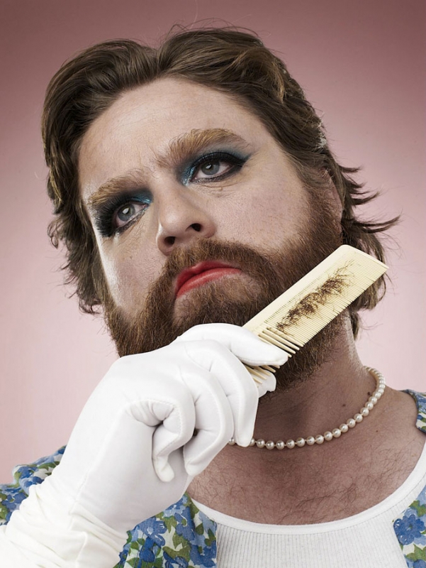 Portrait of a man with a beard wearing makeup, white gloves, pearl necklace, and floral top, combing his beard against a pink background.