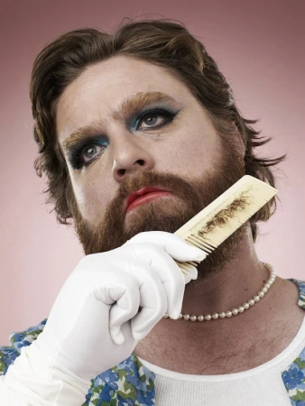 Portrait of a man with a beard wearing makeup, white gloves, pearl necklace, and floral top, combing his beard against a pink background.