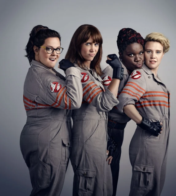Four women in 'Ghostbusters' jumpsuits posing in a photo studio, smiling and looking at the camera with a grey background.