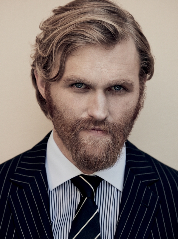 Close-up portrait of a man with a beard and mustache wearing a dark pinstripe suit, white shirt, and striped tie against a neutral background.