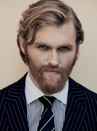 Close-up portrait of a man with a beard and mustache wearing a dark pinstripe suit, white shirt, and striped tie against a neutral background.