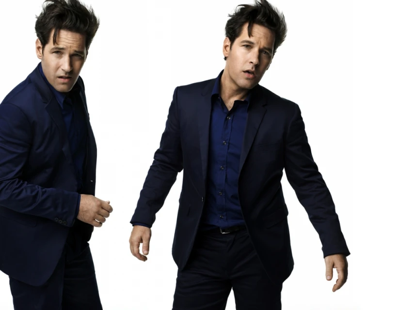 Studio portrait of Paul Rudd wearing a navy suit and blue shirt, posing against a white background, duplicated in the frame.