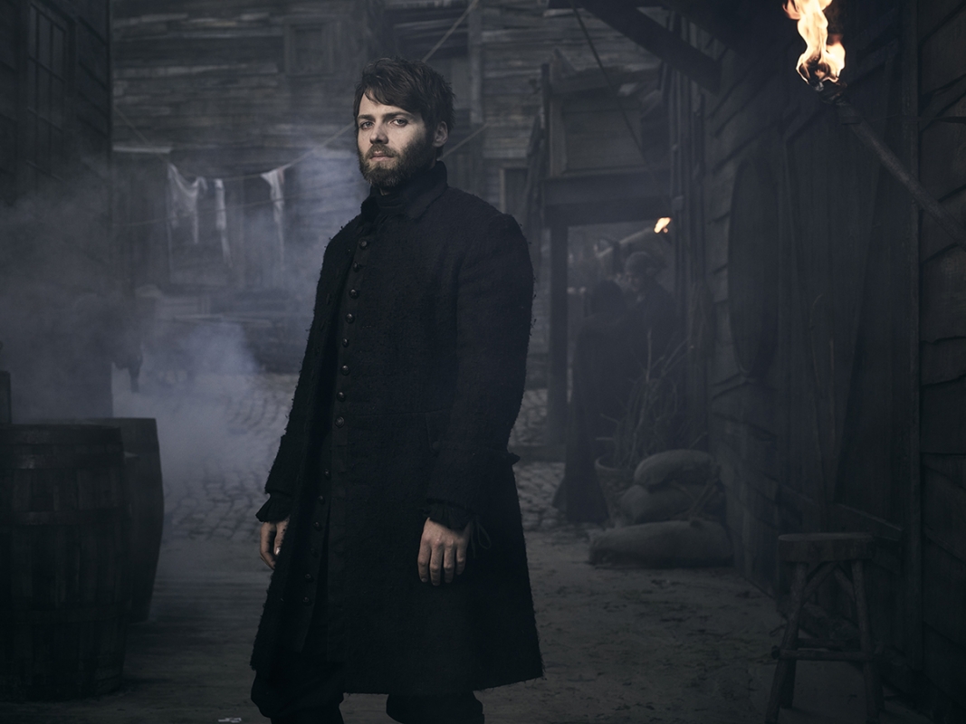 A man in a long black coat stands in a dimly lit town street with buildings, torches, and smoke adding to the atmosphere of a film set.