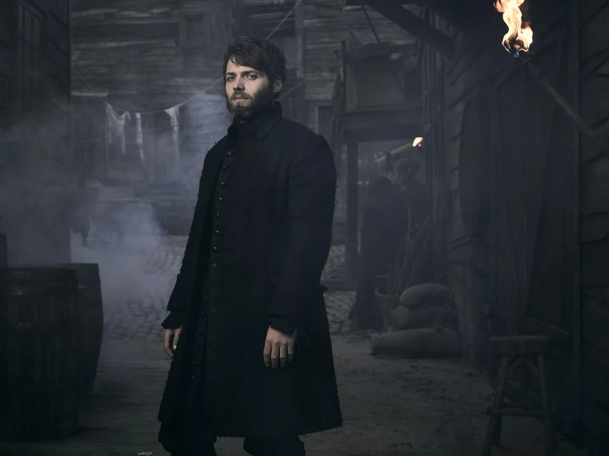 A man in a long black coat stands in a dimly lit town street with buildings, torches, and smoke adding to the atmosphere of a film set.