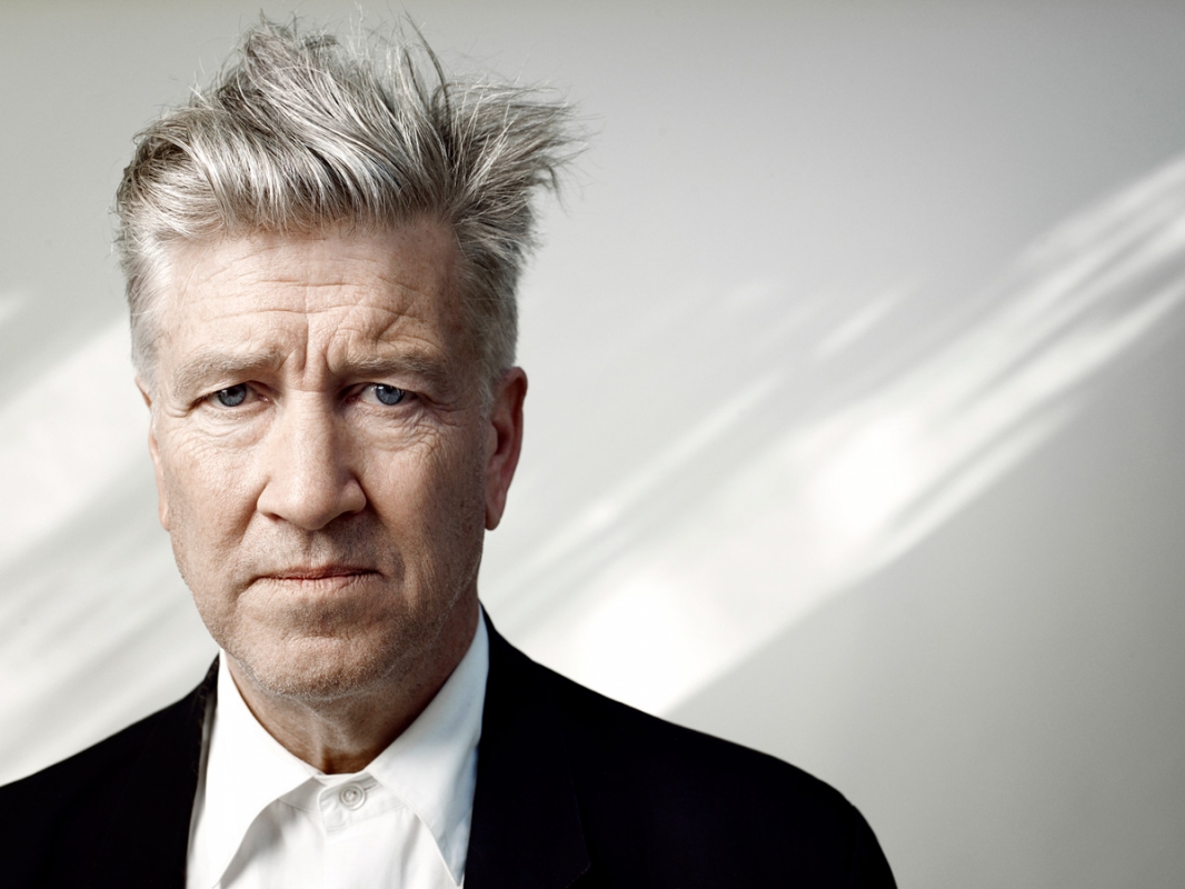 Portrait of David Lynch with his signature gray hair and a black suit jacket against a softly lit background, focusing on his intense gaze.