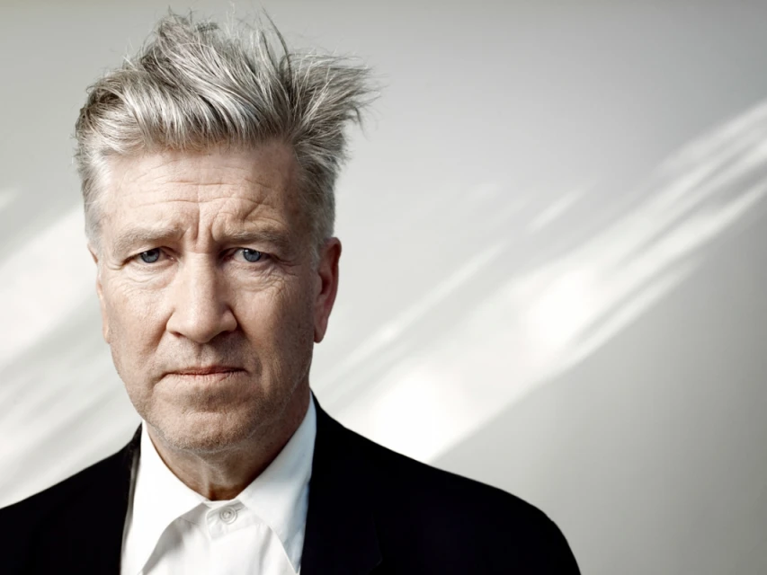 Portrait of David Lynch with his signature gray hair and a black suit jacket against a softly lit background, focusing on his intense gaze.