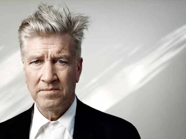 Portrait of David Lynch with his signature gray hair and a black suit jacket against a softly lit background, focusing on his intense gaze.