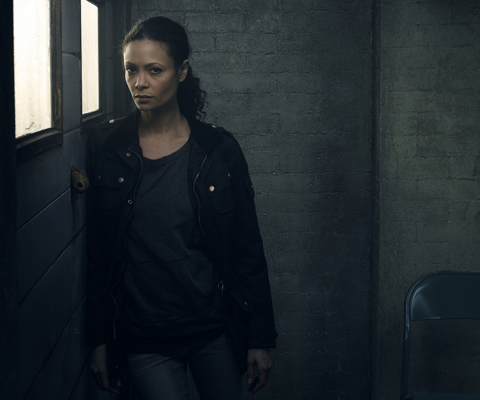 Portrait of an adult woman indoors, leaning against a wall with a serious expression, wearing a dark jacket and a grey shirt.
