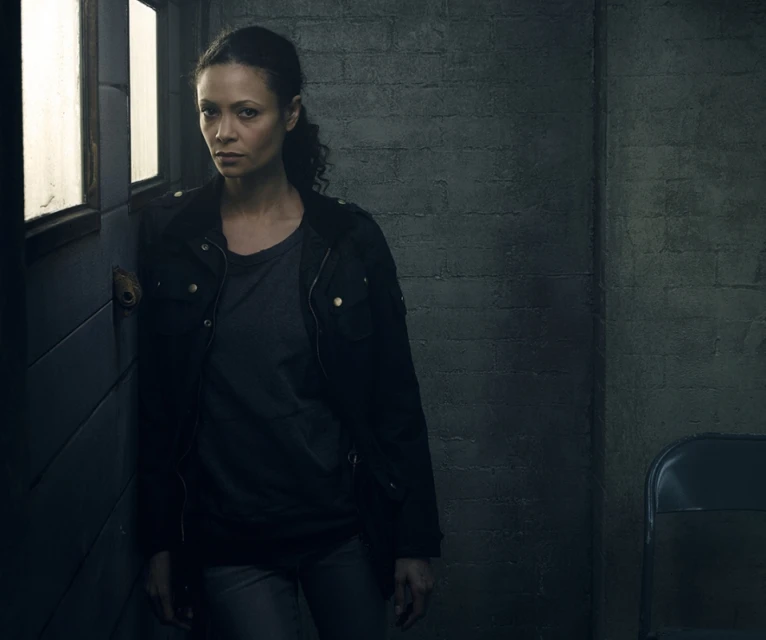 Portrait of an adult woman indoors, leaning against a wall with a serious expression, wearing a dark jacket and a grey shirt.