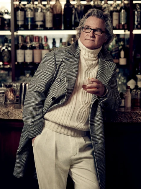 Stylish mature man with grey hair wearing a turtleneck and plaid coat poses indoors with a drink at a bar under warm lighting.