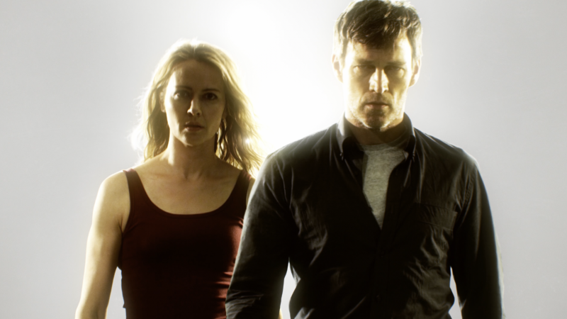 Portrait of a man and woman standing close together in a bright, neutral background, both gazing forward with a serious expression.