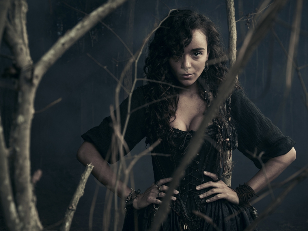 Portrait of a woman in a dark dress standing in front of bare branches with a dark background. She has dark curly hair and fair skin.