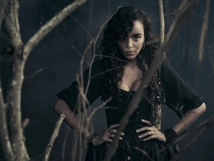 Portrait of a woman in a dark dress standing in front of bare branches with a dark background. She has dark curly hair and fair skin.