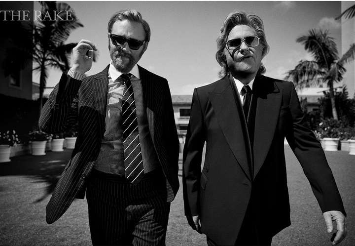 Two adult males in suits and sunglasses stand outdoors on a sunny day. One man smokes a cigarette, while palm trees grow in the background.