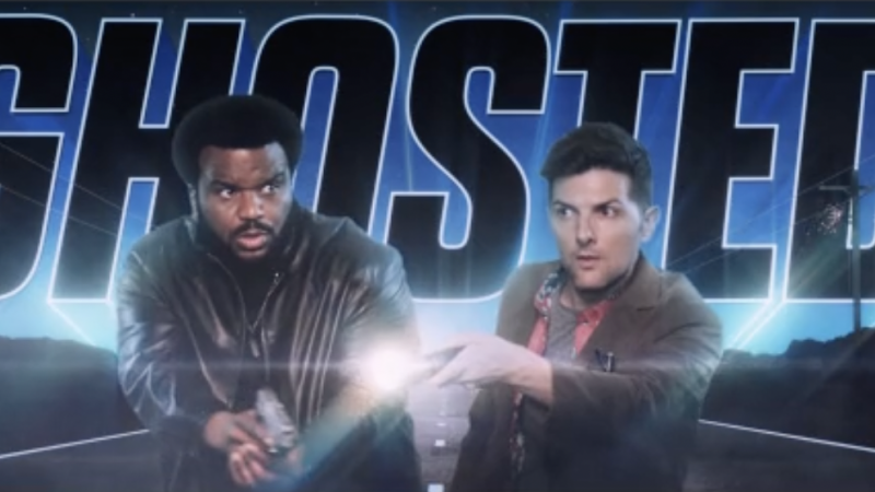 Two men hold guns in front of a promotional backdrop for the film/TV show Ghosted. They wear jackets and look surprised.
