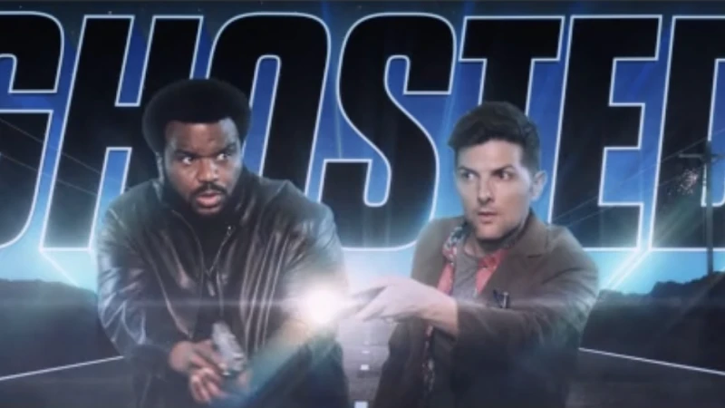 Two men hold guns in front of a promotional backdrop for the film/TV show Ghosted. They wear jackets and look surprised.