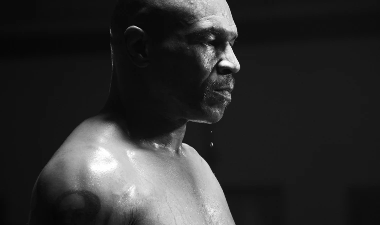 Black and white portrait of a sweaty adult male with a tattoo on his shoulder and a shaved head, dripping water from his chin.