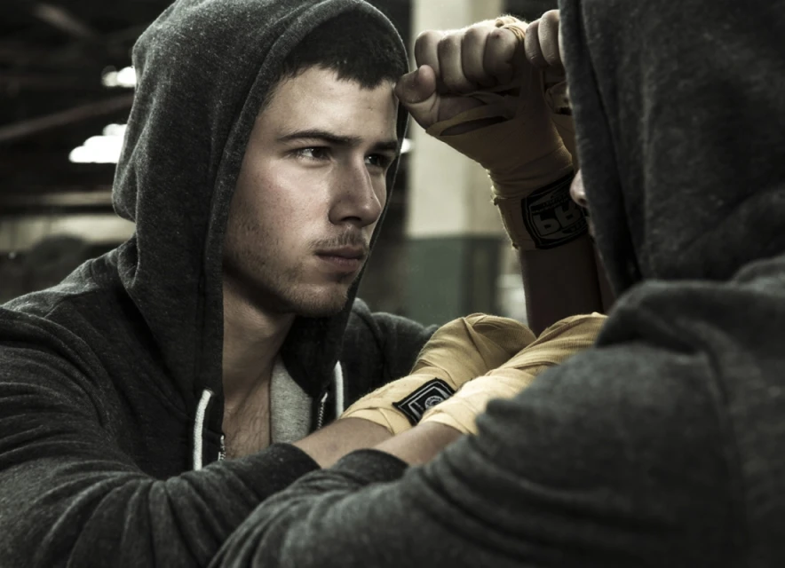 Two men with wrapped hands in a boxing gym, wearing hooded sweatshirts, staring intently in a low-key shot. 