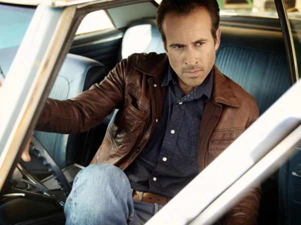 Man wearing a brown leather jacket and blue shirt sitting inside a vintage blue car