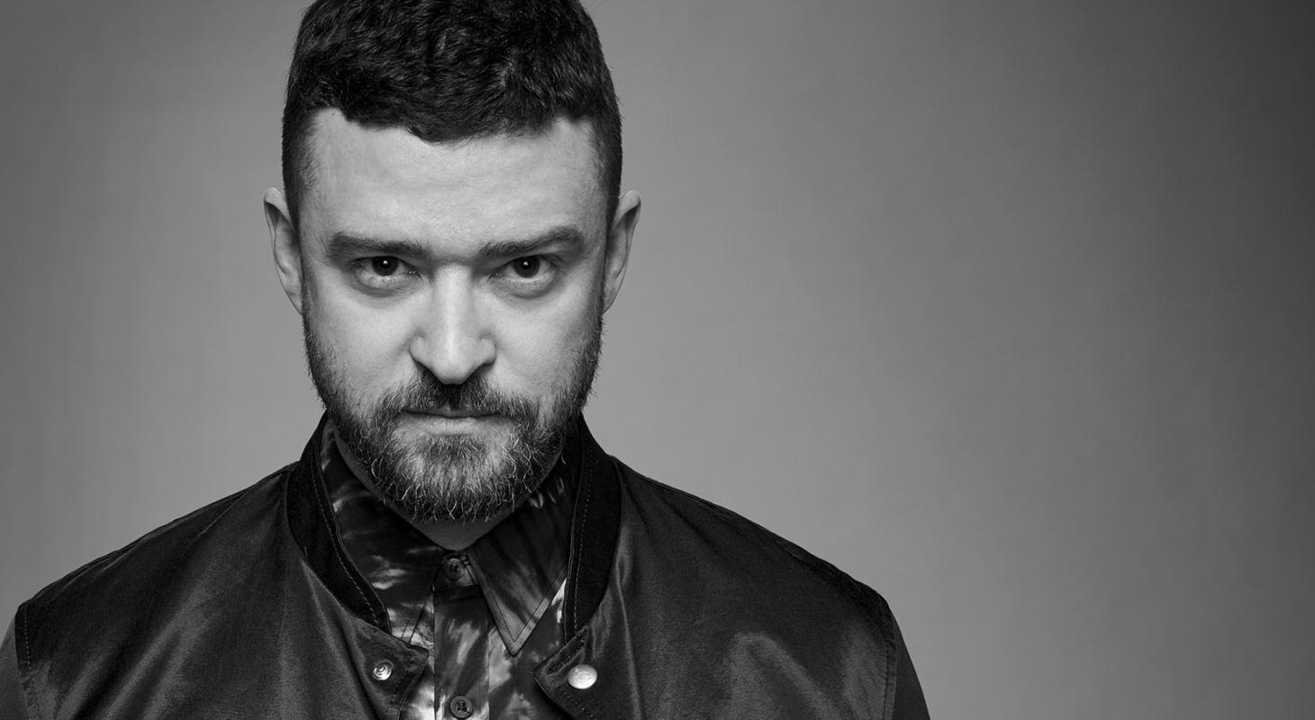 Black and white portrait of Justin Timberlake looking at the camera with a serious expression in a collared shirt and bomber jacket.
