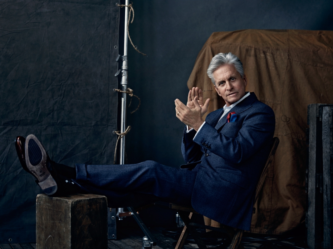 Michael Douglas poses in a blue suit indoors with hands together, wearing dress shoes, seated with lighting equipment visible in the background.
