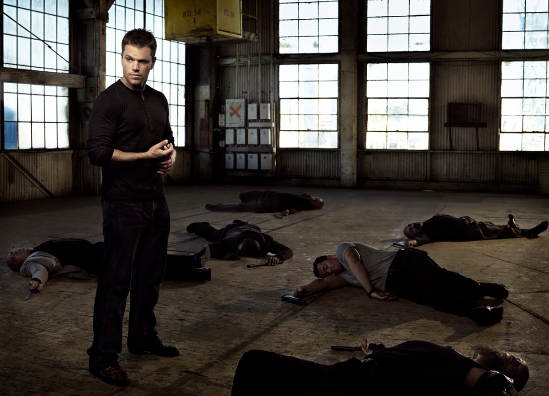 Matt Damon standing in a dimly lit industrial warehouse filled with several fallen figures on the concrete floor.