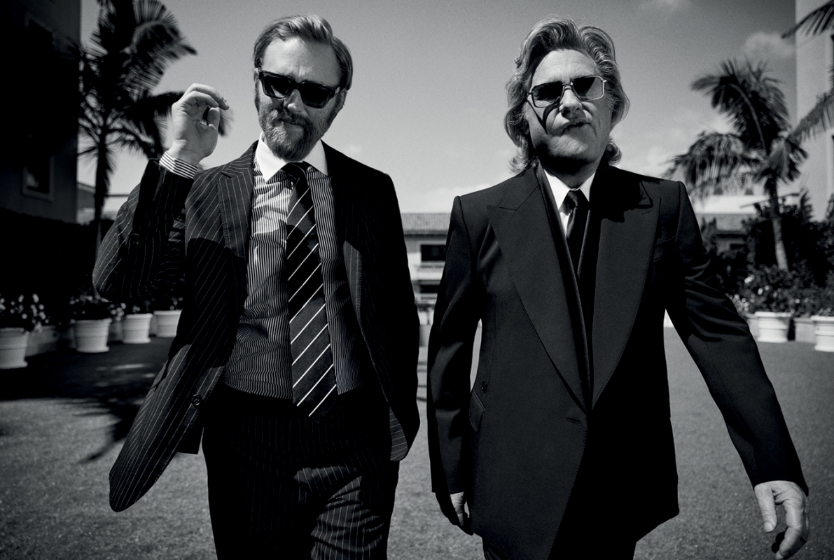 Two men in dark suits and sunglasses walking outdoors near palm trees in a monochrome portrait by Justin Stephens