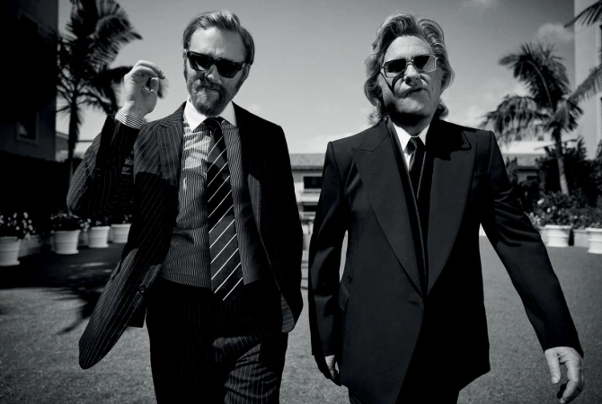 Two men in dark suits and sunglasses walking outdoors near palm trees in a monochrome portrait by Justin Stephens