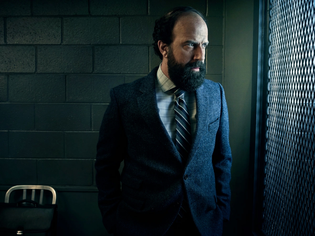Man in a suit stands next to a metal structure in an indoor setting. He has dark hair, a beard, and a serious expression.