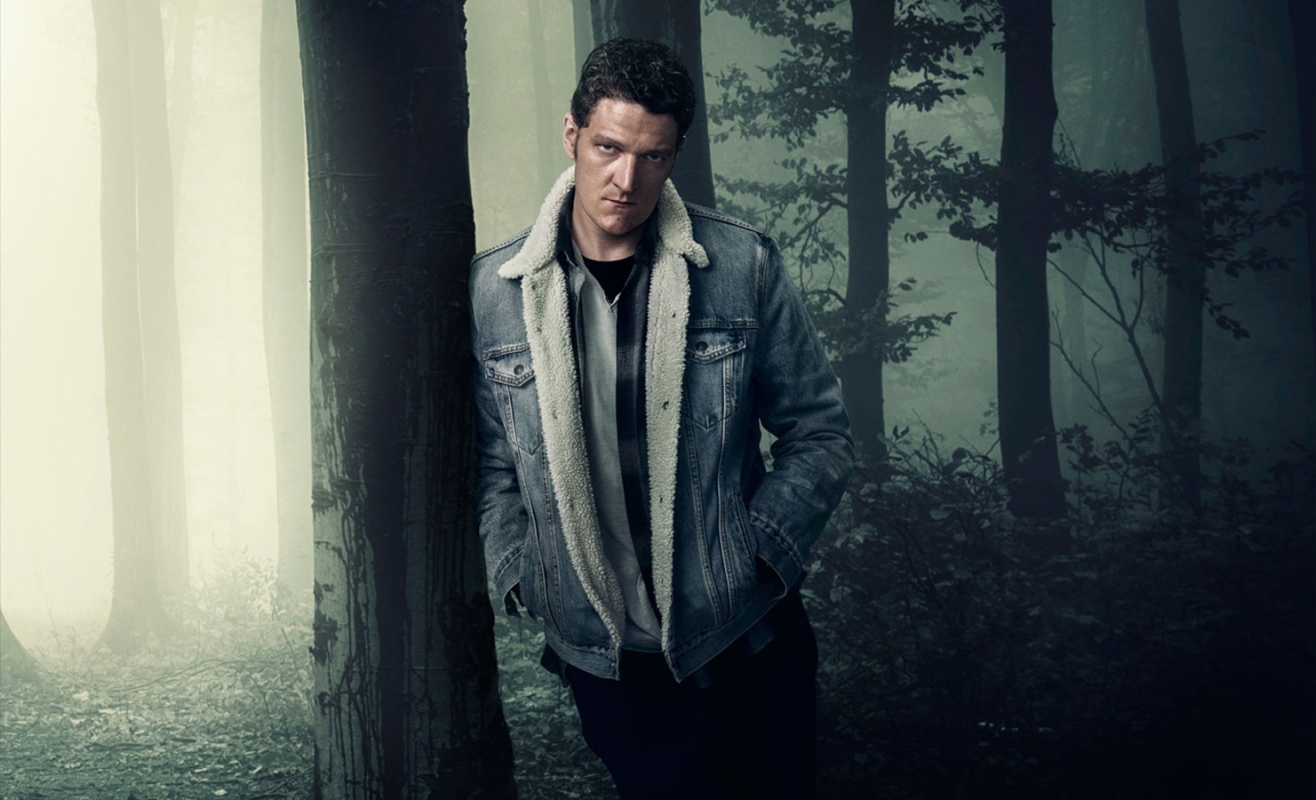Portrait of a man in a denim jacket with a fur collar, leaning against a tree in a foggy forest, looking intensely at the camera.