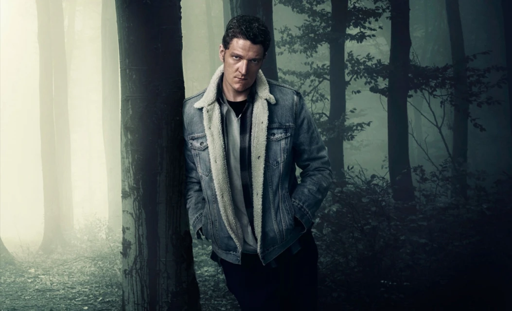 Portrait of a man in a denim jacket with a fur collar, leaning against a tree in a foggy forest, looking intensely at the camera.