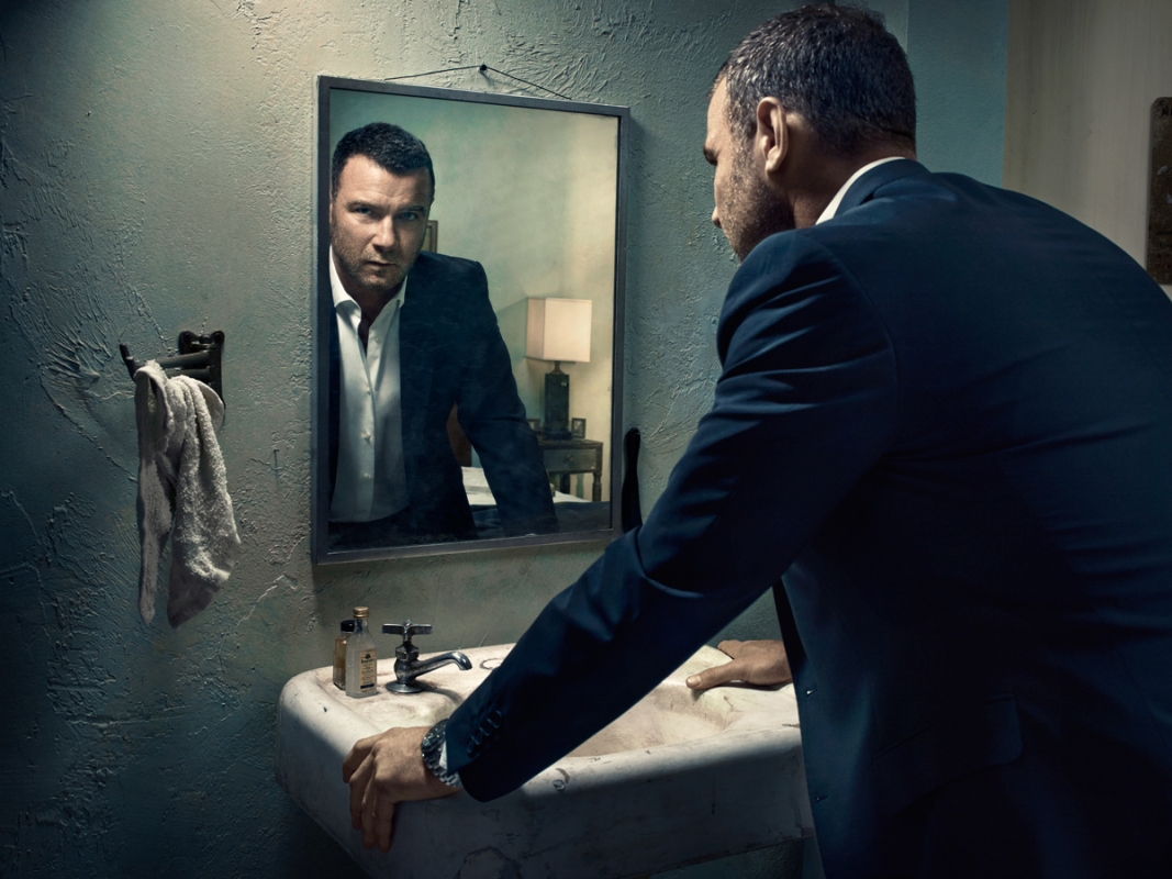 Man in a suit looks at his reflection in a dimly lit bathroom with a white sink, mirror, and textured walls and towel rack.