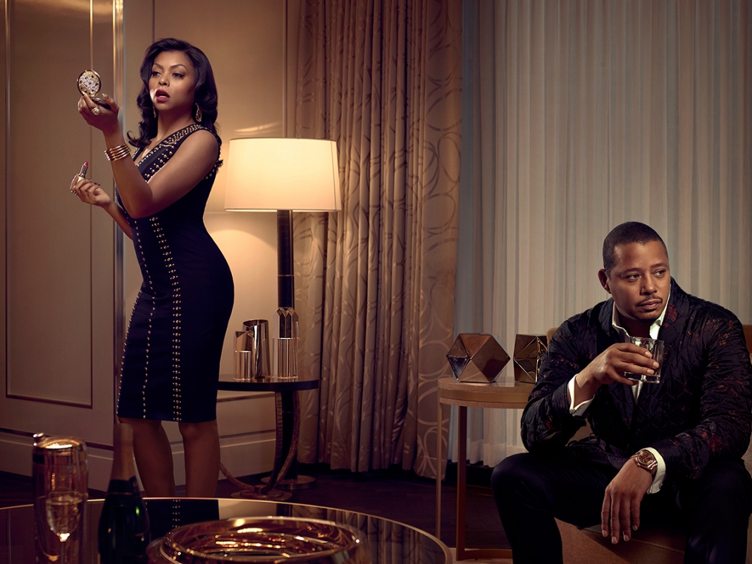 Glamorous woman in black dress applying lipstick, man seated in the background in a modern hotel room.