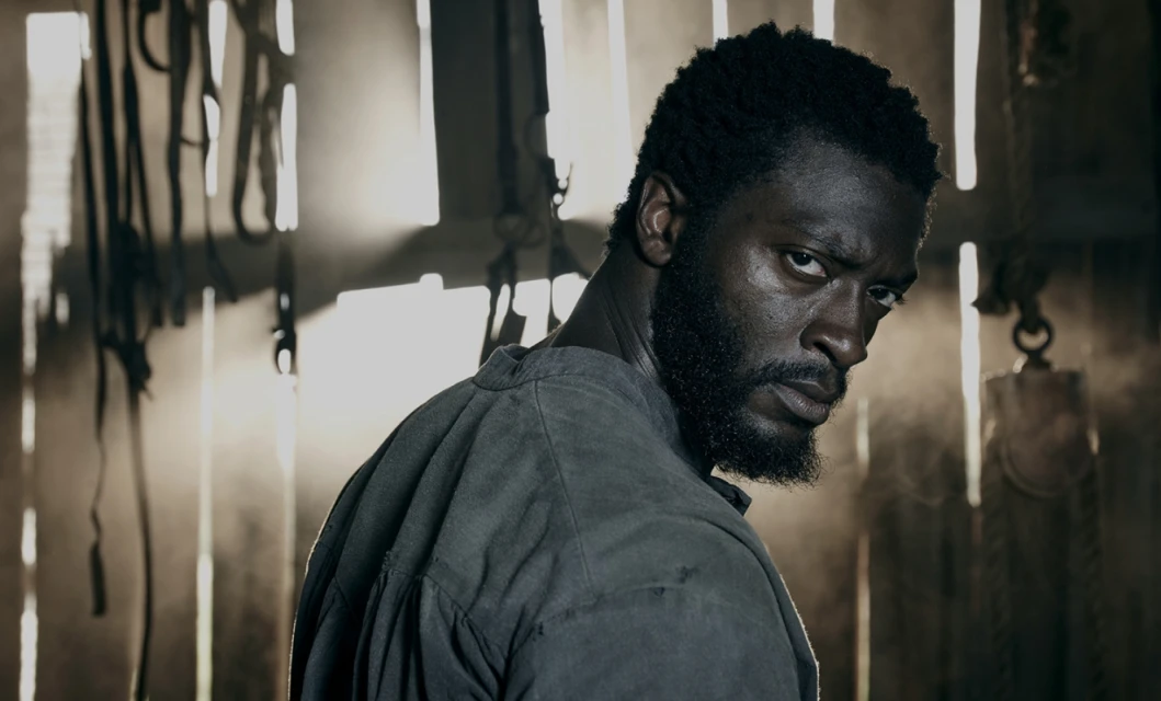 Serious-looking African-American man gazes intensely over his shoulder in a dimly lit, rustic indoor environment with visible woodwork.