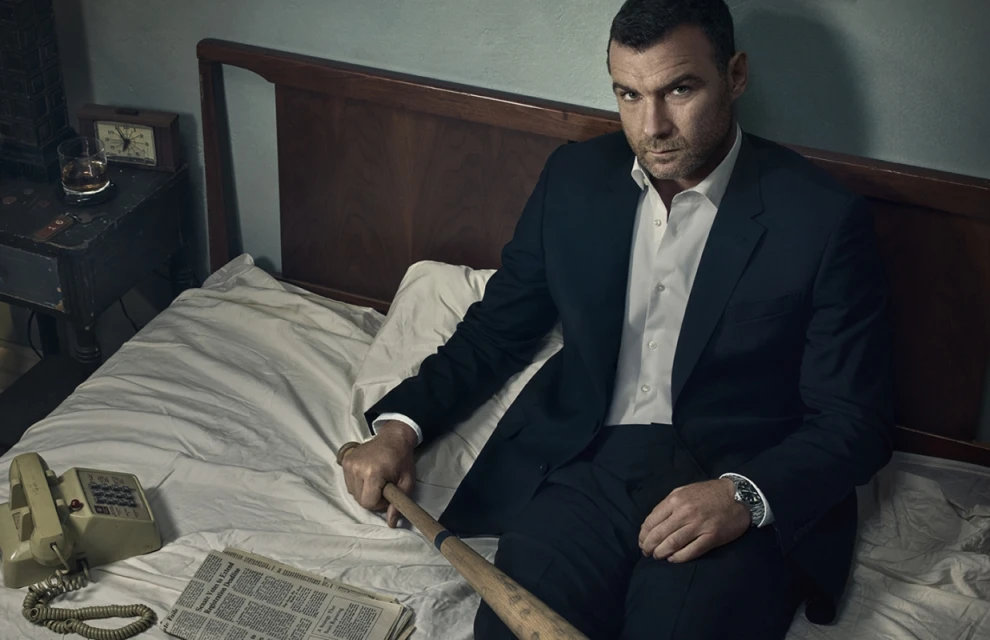 Adult male in a dark suit sits on a bed holding a baseball bat with a newspaper and vintage phone beside him in an indoor setting.