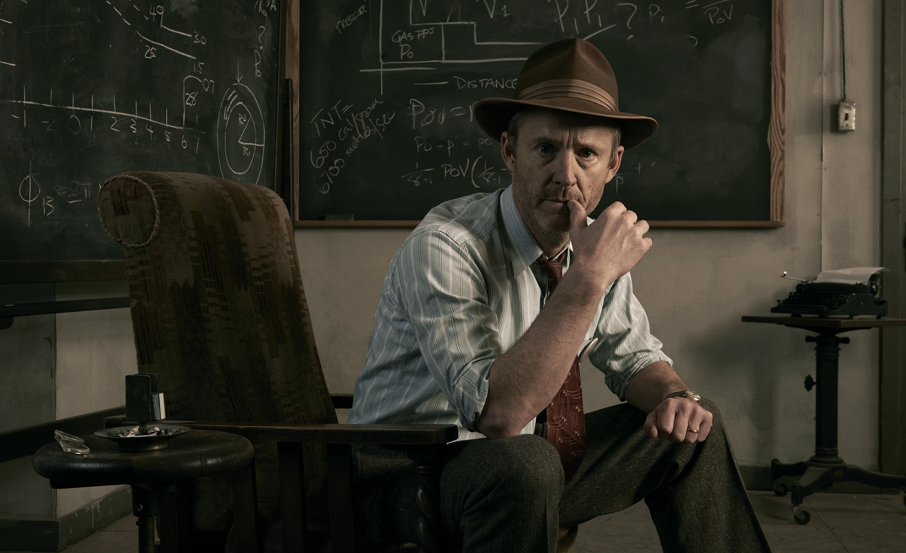 Man in a hat sits thoughtfully in a striped armchair in an office, with chalk equations on a blackboard behind him and a typewriter visible.