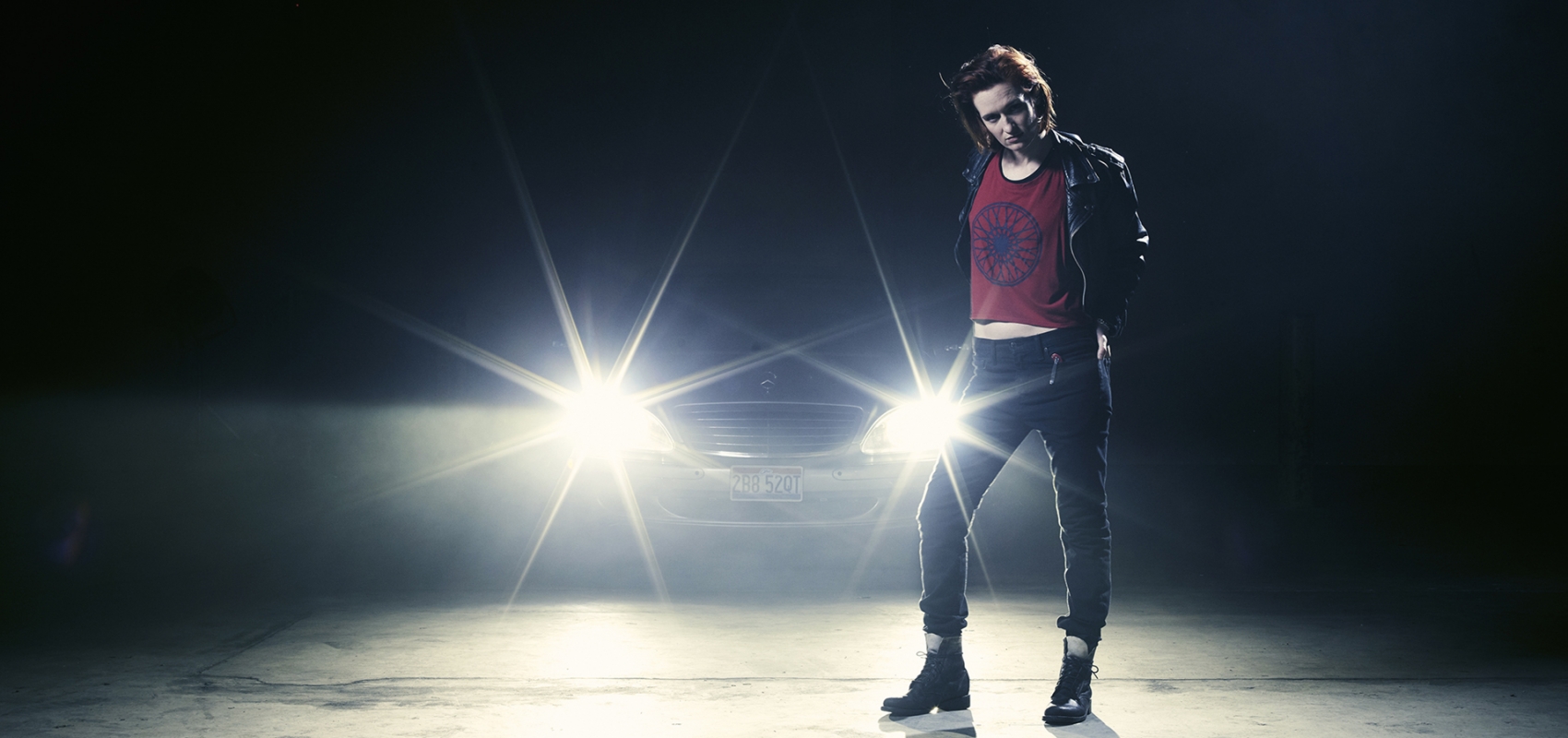 Woman in black leather jacket and jeans poses in front of a car with headlights beaming, creating a dramatic and edgy studio portrait.