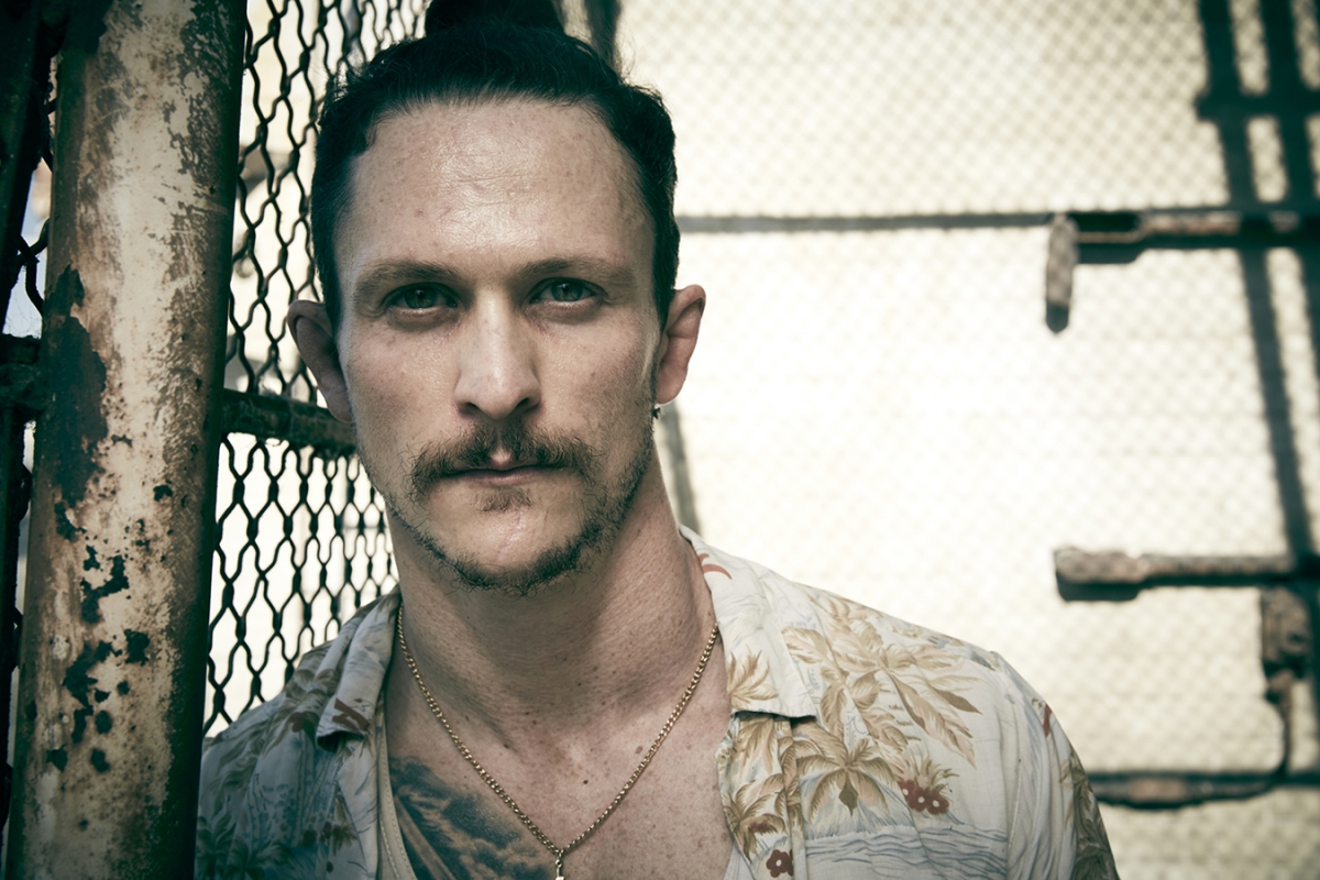 Close-up portrait of a man with a mustache and a topknot, wearing a tropical print shirt, standing against a metal gate with a neutral expression.
