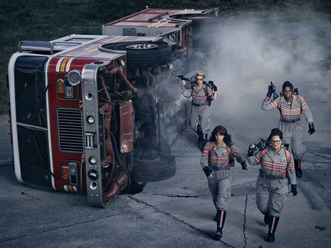 Four women in grey jumpsuits walk toward an overturned red and white fire truck on a concrete road with green trees in the distance.
