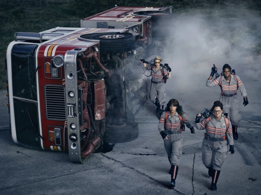 Four women in grey jumpsuits walk toward an overturned red and white fire truck on a concrete road with green trees in the distance.