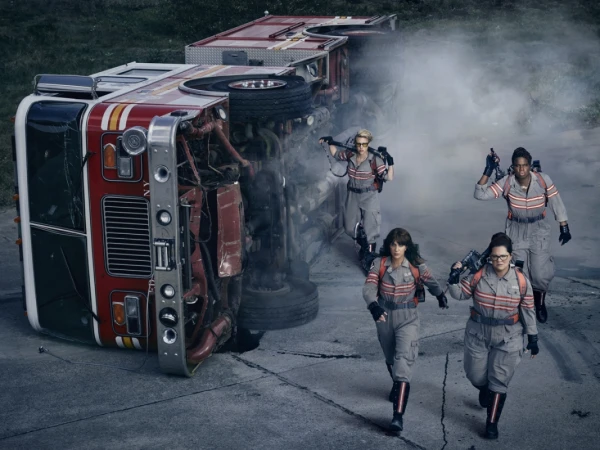 Four women in grey jumpsuits walk toward an overturned red and white fire truck on a concrete road with green trees in the distance.