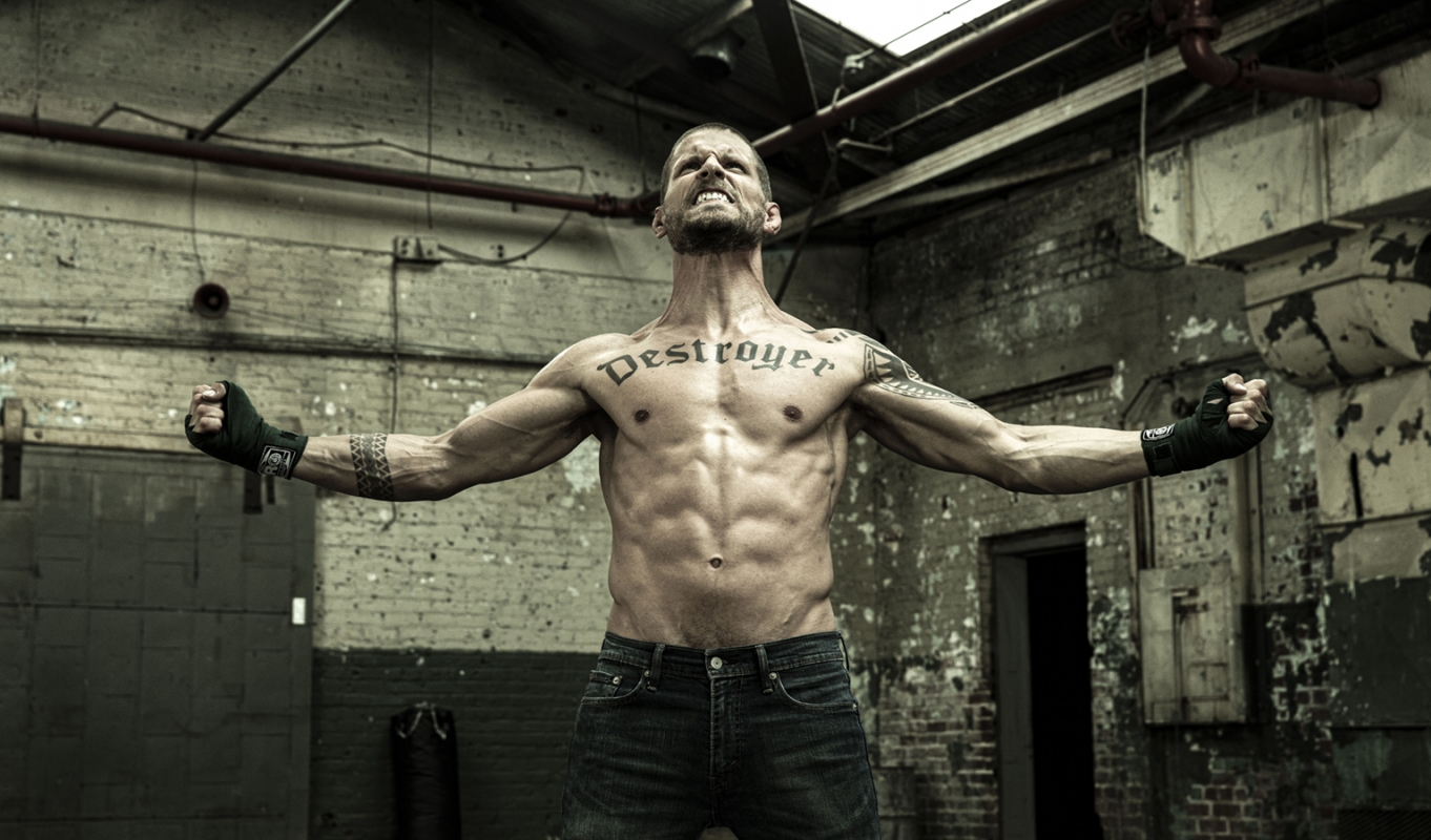 Muscular man flexing in a gritty industrial setting with exposed brick and pipes, wearing jeans and wrist wraps, showcasing chest tattoo.
