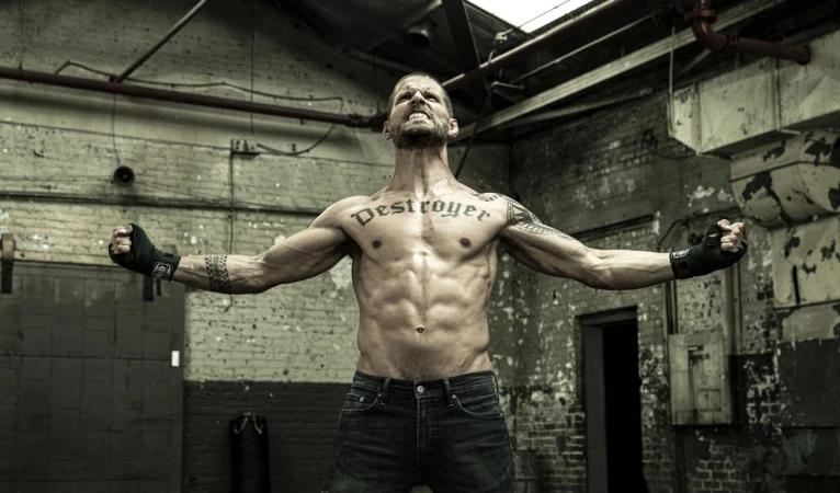 Muscular man flexing in a gritty industrial setting with exposed brick and pipes, wearing jeans and wrist wraps, showcasing chest tattoo.