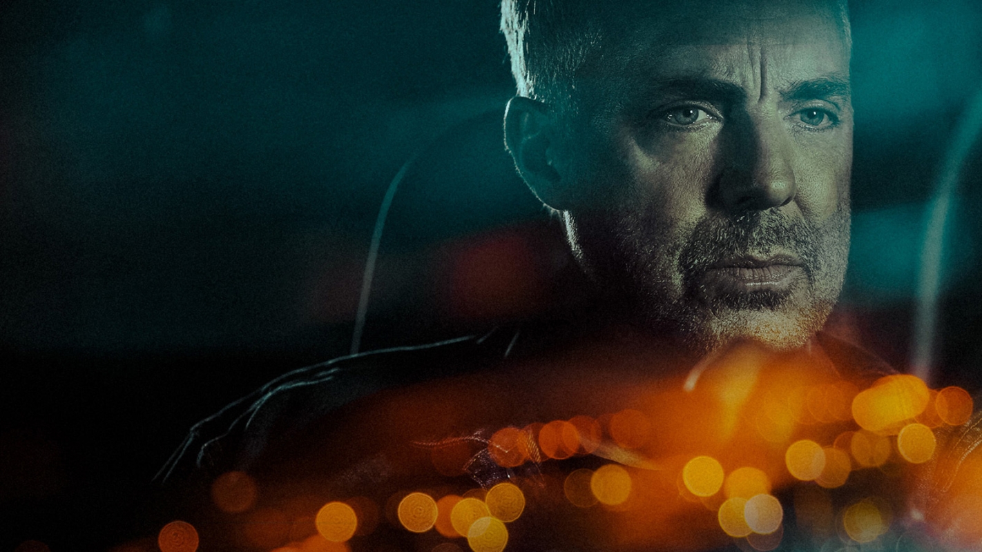 Portrait of a man with a serious expression, partially obscured by out-of-focus orange lights against a dark teal background.